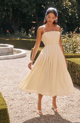 A woman is wearing a cream-colored, pleated midi dress with thin spaghetti straps. She has a multicolored headscarf tied around her head. She is also wearing white high-heeled sandals with an open toe. The background features a garden with greenery, flowers, and a small round fountain.